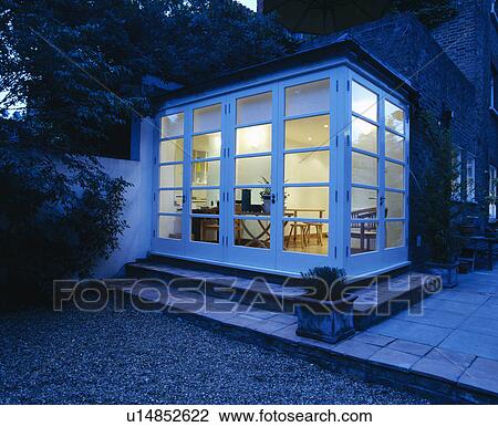 Stock Image - View from outside through windows of modern conservatory extension at early evening with lights on inside. Fotosearch
