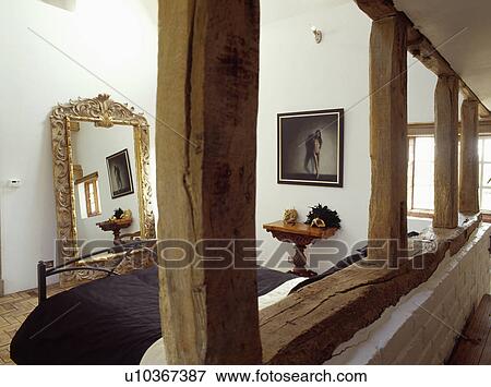 View through large beams of wall with plaster removed of white attic bedroom with large gilt mirror on floor View Large Photo Image Stock Photo - View through large beams of wall with plaster removed of white attic bedroom with large gilt mirror on floor. Fotosearch