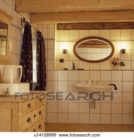 Stock Photo - Wall-lights and oval mirror above wall-mounted basin in white tiled bathroom with beamed ceiling and pine chest of drawers. Fotosearch