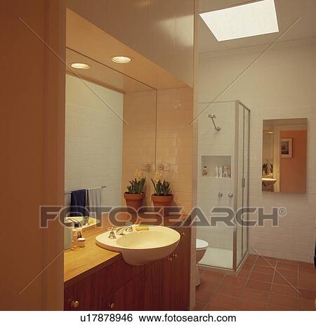 Stock Photograph - White basin set into pale wood vanity unit below mirror in modern white bathroom with terracotta tiled floor . Fotosearch