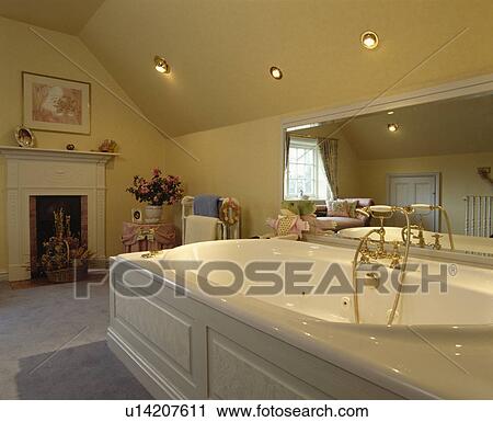 Stock Image - White bath with grey panelling below long mirror in traditional attic bathroom with small fireplace. Fotosearch