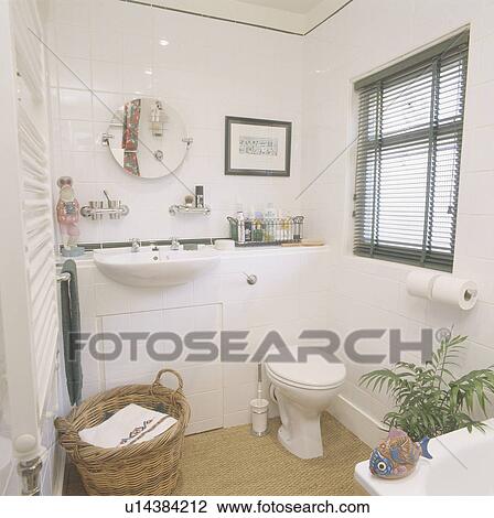 Stock Image - White built in basin in vanity unit in small white bathroom with black slatted blind and sisal carpet & basket of towels. Fotosearch