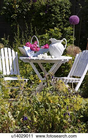 Stock Image - White chairs and table with white jug in country garden in summer. Fotosearch