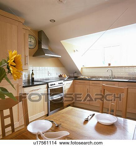 Picture - White crockery on plain wood table in modern attic kitchen with pale wood fitted cupboards. Fotosearch