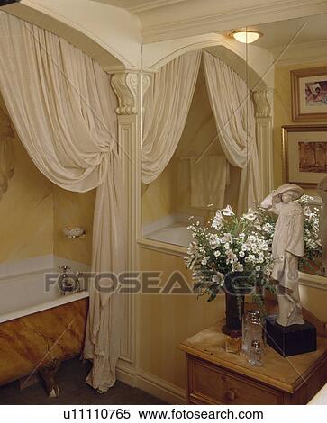 Stock Photography - White flowers and Edwardian statue on table in front of mirrored wall in bathroom with cream curtain beside bath. Fotosearch