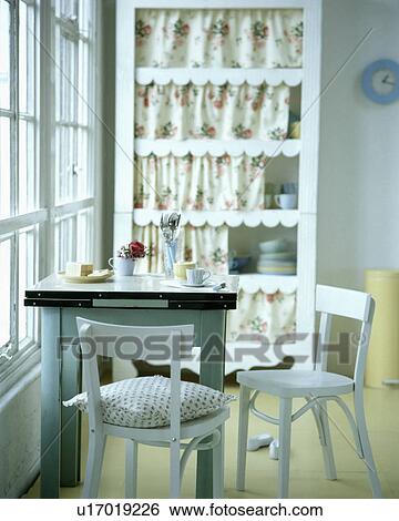 Stock Photograph - White forties chairs and zinc-topped table in white dining room with yellow floor and cupboard with floral curtains. Fotosearch