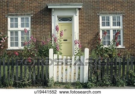 Stock Image - White gate in wooden fence in front of cottage with white windows and yellow front door & foxgloves in the garden. Fotosearch