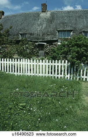 Stock Image - White picket fence in front of thatched country cottage. Fotosearch