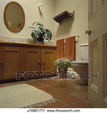 Stock Photo - White rug on terracotta floor tiles in bathroom with houseplants and wooden vanity unit. Fotosearch