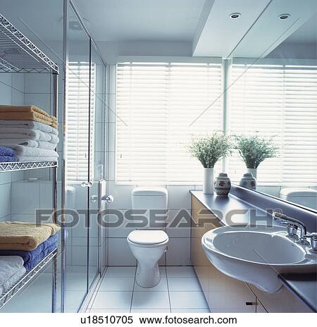Stock Photography - White slatted blind above toilet in small modern white mirrored bathroom with towels on stainless steel shelving. Fotosearch