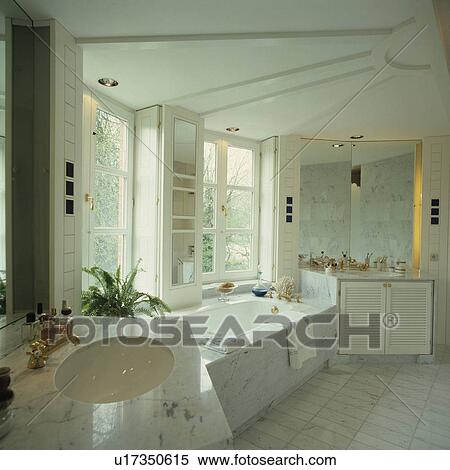 Stock Photography - White underset basin in marble vanity unit in white bathroom with marble surround on bath below windows. Fotosearch