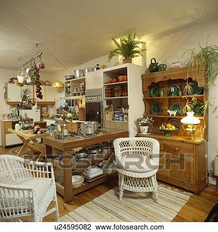 Stock Image - White wicker chairs and pine dresser with lighted lamp in large traditional kitchen. Fotosearch