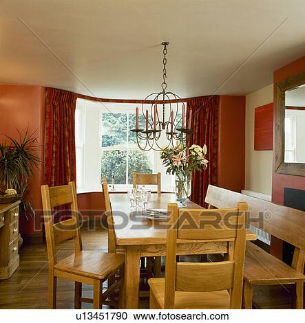 Stock Image - Wooden chairs and bench at wooden table below metal candelabra in terracotta diningroom. Fotosearch