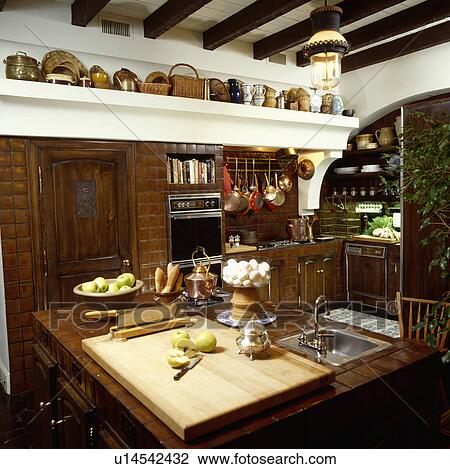 Stock Image - Wooden chopping board and steel sink in island unit in traditional kitchen. Fotosearch