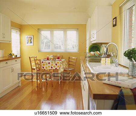 Wooden flooring in modern yellow kitchen with fitted white cupboards and rush-seated chairs at table with spotted cloth View Large Photo Image Stock Photo - Wooden flooring in modern yellow kitchen with fitted white cupboards and rush-seated chairs at table with spotted cloth. Fotosearch