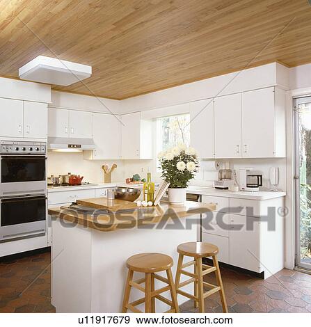 Stock Photo - Wooden stools and island unit with wood worktop in modern white kitchen with wooden ceiling. Fotosearch