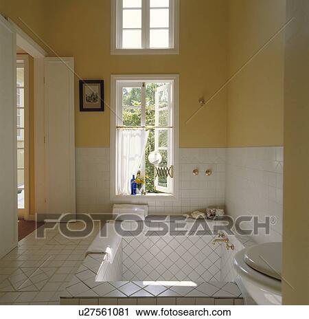 Stock Image - Yellow painted, white tiled bathroom with large sunken bath. Fotosearch