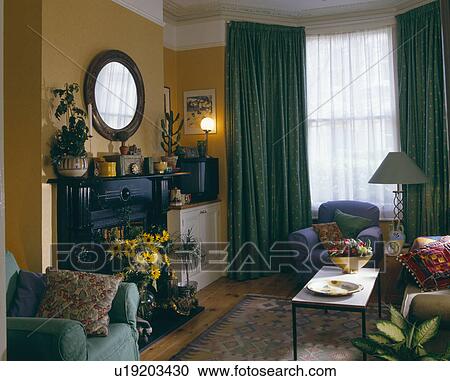 白 網 そして 緑 カーテン 中に 小さい Livingroom で 黄色の壁 そして オバール 鏡 の上 暖炉 ストックイメージ U Fotosearch