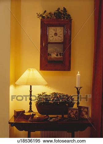 Stock Photography - Antique clock with painted scene above lighted lamp on small table in country hall. Fotosearch