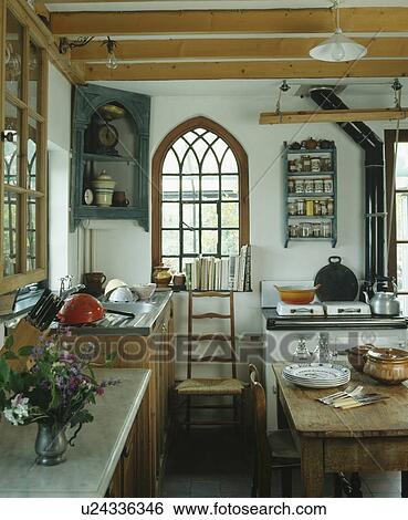 Stock Photograph - Antique corner cupboard beside arched Gothic window above rush-seated antique chair in country kitchen. Fotosearch