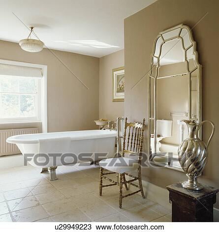 Antique Gilt Chair In Front Of Large Mirror Neutral Bathroom With White Rolltop Bath And Limestone Floor Tiles Stock Image