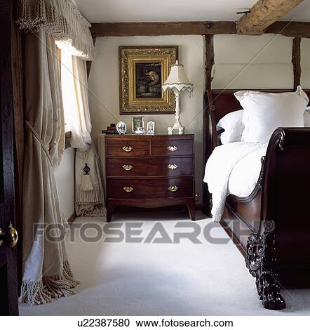 Stock Image - Antique mahogany chest-of-drawers and lit bateau bed in cottage bedroom with white carpet and cream curtains. Fotosearch