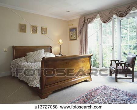 Stock Photo - Antique mahogany lit bateau bed in bedroom with grey swagged curtains at French windows. Fotosearch