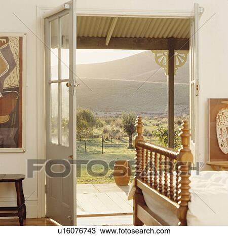 Stock Image - Antique wooden bed with turned spindles in white Australian bedroom with view of countryside through open door. Fotosearch