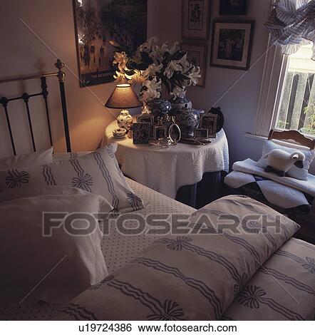 Stock Photograph - Bed with blue+white ribbon+bow patterned duvet and pillows beside bedside table with lighted lamp and photographs. Fotosearch