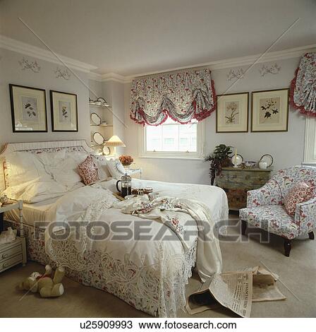 Stock Image - Bed with lace bedcover in bedroom with floral festoon blinds and matching armchair. Fotosearch