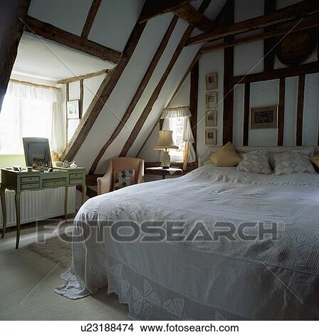 Picture - Bed with lace bedspread in beamed attic bedroom with dressingtable in front of dormer window. Fotosearch