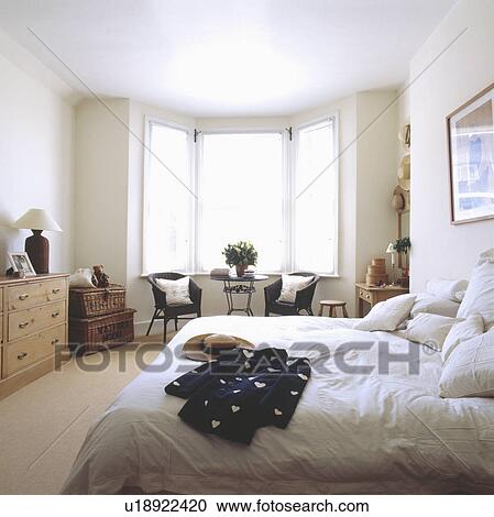 Stock Image - Black & white jacket on bed with white bedlinen in white bedroom with white blinds and wicker chests. Fotosearch