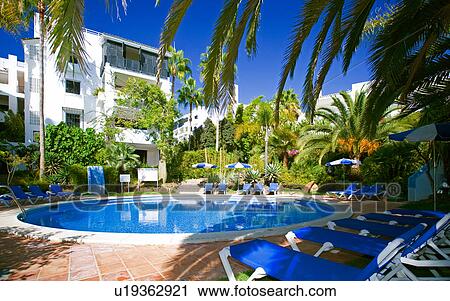 Stock Image - Blue chaiselongues around blue circular swimmingpool in front of modern white Spanish apartment building. Fotosearch