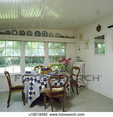 Stock Image - Blue+white patchwork cloth on table in front of window white country dining room with white timbered ceiling and walls . Fotosearch