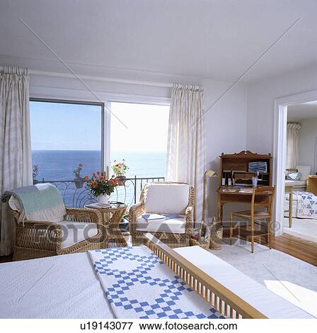 Blue+white patchwork quilt on bed in white bedroom with wicker armchairs in front of windows with view of the ocean View Large Photo Image Stock Photo - Blue+white patchwork quilt on bed in white bedroom with wicker armchairs in front of windows with view of the ocean. Fotosearch
