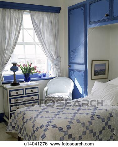 Stock Photograph - Blue+white patchwork quilt on bed in alcove with fitted blue cupboards in bedroom with white curtains. Fotosearch