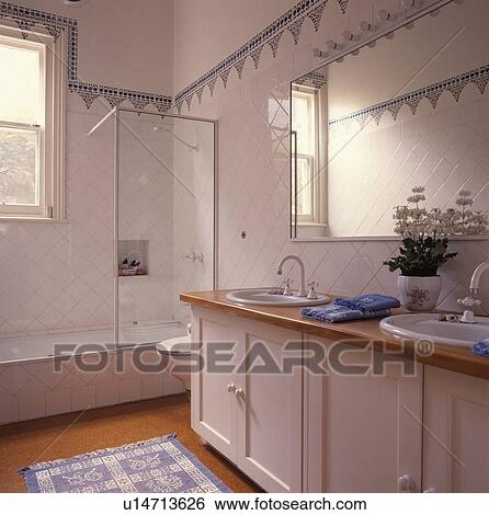 Stock Photograph - Blue+white tiled border above white tiled walls in white bathroom with double basins in traditional white vanity unit. Fotosearch