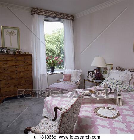 Stock Photography - Breakfast tray and pink patchwork quilt on bed in traditional white country bedroom with white voile curtains on window. Fotosearch