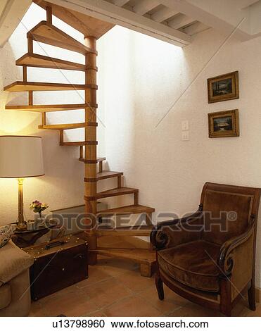 Stock Image - Brown antique armchair below wooden spiral staircase in living room with lighted lamp. Fotosearch