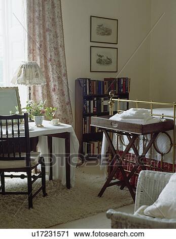 Stock Image - Butler's Tray at the foot of brass bed and white cloth on table in front of the window in country bedroom. Fotosearch