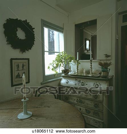 Stock Image - Candle on circular pine table in small dining room with floral painted bureau beside window. Fotosearch