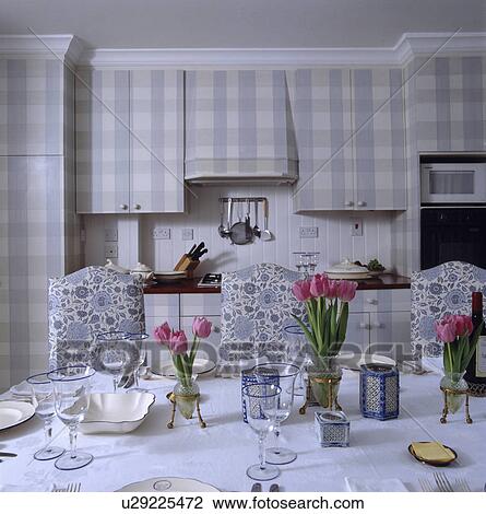 Stock Image - China and glasses on table with white cloth and blue floral patterned chairs in kitchen with blue+white check cupboards. Fotosearch