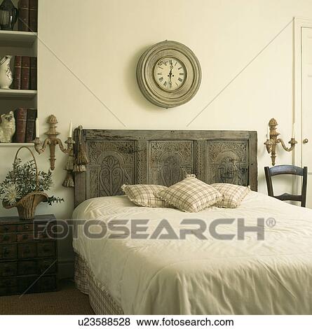 Circular distressed wooden clock above bed with reclaimed carved antique headboard in bedroom with cream bedlinen View Large Photo Image Stock Photo - Circular distressed wooden clock above bed with reclaimed carved antique headboard in bedroom with cream bedlinen. Fotosearch