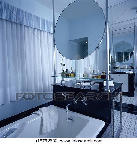 Stock Image - Circular mirror above black unit with stainless steel tap pouring water into white bath in modern bathroom. Fotosearch