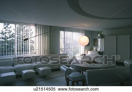 Stock Photography - Circular stone table and white leather seating in white dining and living room with white sofa in front of large window. Fotosearch