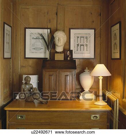 Stock Image - Classical bust and urn on chest of drawers with small statue and lighted lamp in panelled alcove. Fotosearch