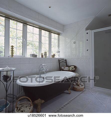 Stock Image - Clawfoot bath below windows in white and grey tiled bathroom. Fotosearch