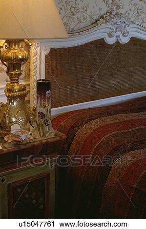 Close-up of gilt lamp on bedside table beside antique French cane bed with dark red and brown bedcover View Large Photo Image Stock Image - Close-up of gilt lamp on bedside table beside antique French cane bed with dark red and brown bedcover. Fotosearch