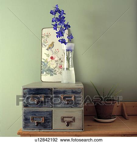 Stock Image - Close-up of oriental picture and blue delphinium in glass vase on miniature drawers. Fotosearch