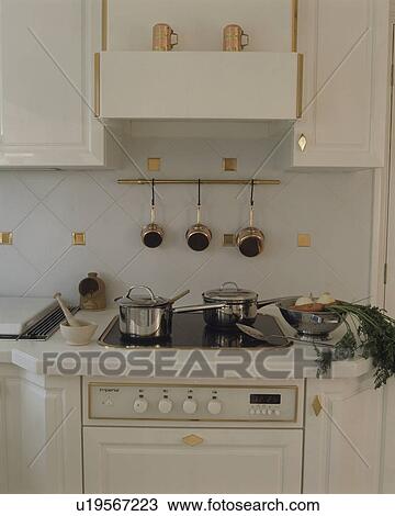 Stock Image - Close-up of stainless steel pans on halogen hob above white oven. Fotosearch